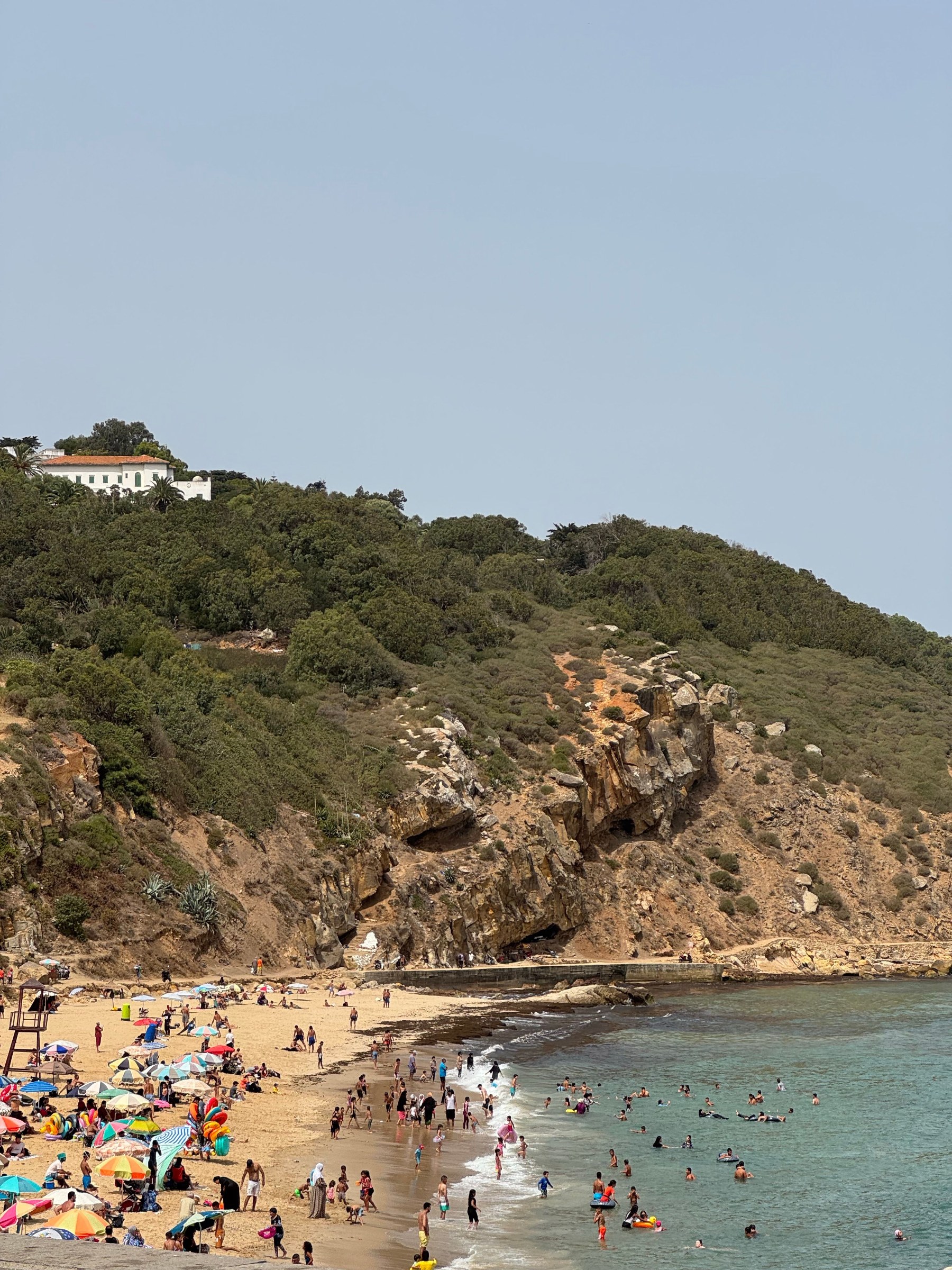 Crowded summer beach below a wooded headland on the Mediterranean coast